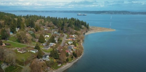 Aerial view of a coastal area with homes, trees, and a distant ship on the water under a cloudy sky.