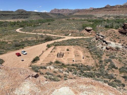Aerial view of a desert landscape with a small garden area, surrounded by rocky terrain and distant hills. Two cars parked nearby.