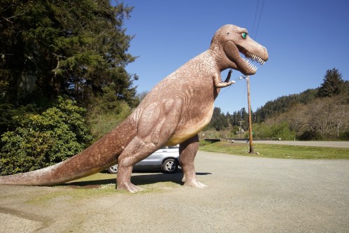 A large dinosaur statue stands beside a parked car in a scenic outdoor setting with trees and blue sky.
