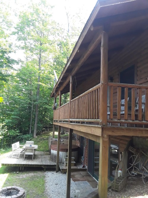 A wooden cabin with a balcony, surrounded by trees, featuring a fire pit and outdoor seating area.