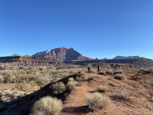 Two people walking on a dirt path with mountains in the background under a clear blue sky.