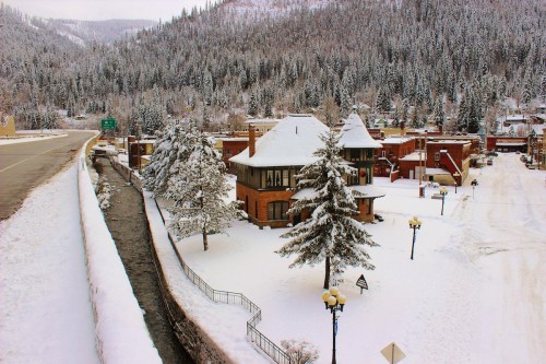 Snow-covered town with historic buildings, pine trees, and a winding creek, surrounded by mountains.