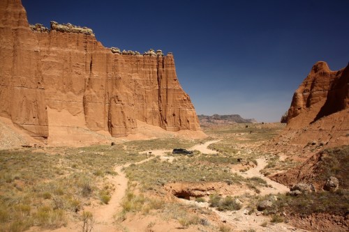 A rugged desert landscape with towering red rock formations and a winding dirt path, under a clear blue sky.