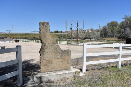 A weathered stone monument shaped like a state, surrounded by a gravel area and white fences under a clear blue sky.