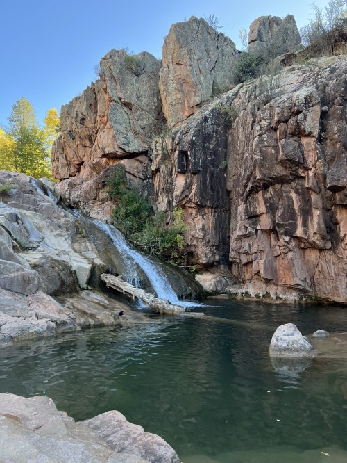 A serene waterfall cascades over rocks into a clear pool, surrounded by rugged cliffs and greenery.