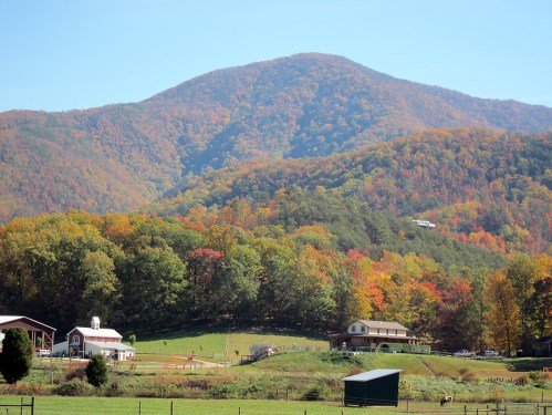 A scenic view of colorful autumn foliage on mountains, with a farm and buildings in the foreground.