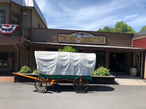 A covered wagon in front of a building labeled "Western Mercantile," with greenery and a clear blue sky in the background.