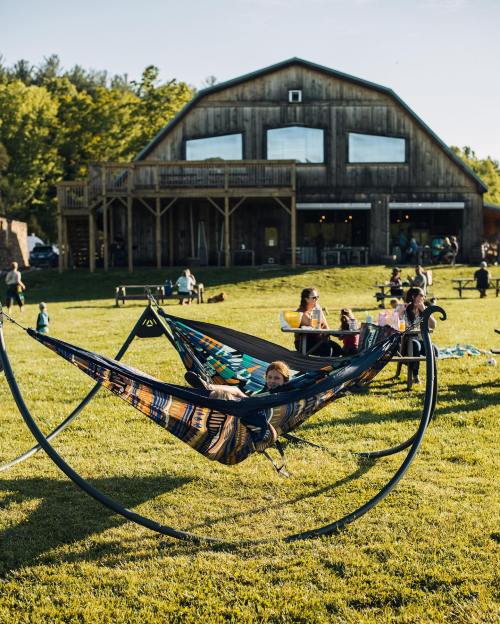 A child relaxes in a colorful hammock outside a rustic barn, with people enjoying the sunny day on the lawn.