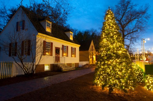 A cozy white house with warm lights beside a brightly lit Christmas tree at dusk.