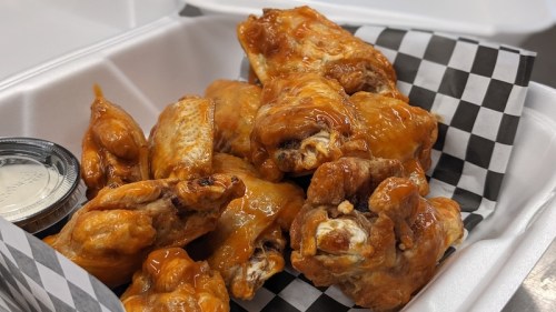A container filled with saucy chicken wings, accompanied by a small cup of dipping sauce and lined with checkered paper.
