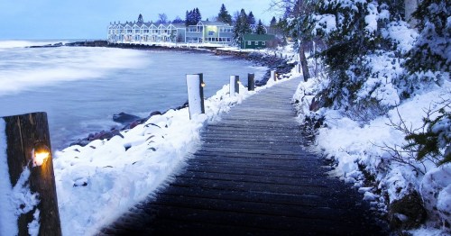 A snowy pathway along the coast, with a view of colorful buildings and a calm sea under a blue sky.