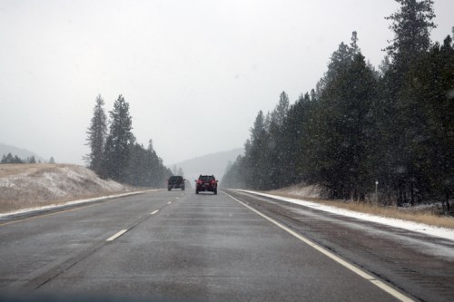 A snowy highway scene with vehicles driving through a winter landscape, surrounded by trees and mountains in the distance.