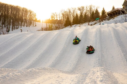 Two people on snow tubes slide down a snowy hill at sunset, surrounded by trees and a winter landscape.