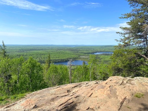A scenic view from a rocky outcrop overlooking lush green forests and a tranquil lake under a clear blue sky.