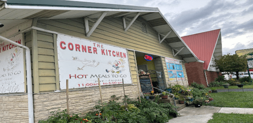 Exterior of The Corner Kitchen, featuring a sign for hot meals to go and an "Open" sign, with plants outside.
