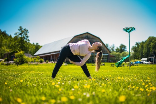 A woman performs a yoga pose in a sunny field with yellow flowers, a barn, and trees in the background.