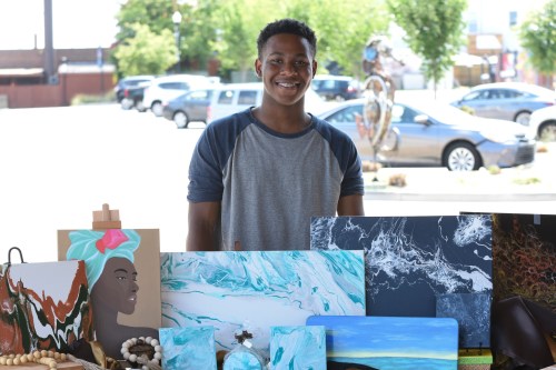 A smiling young man stands behind a display of colorful artwork and crafts at an outdoor market.