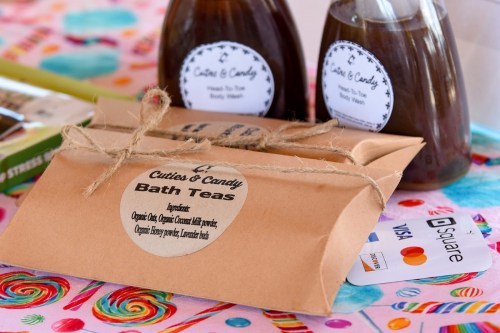 A package of "Cuties & Candy Bath Teas" with labels, surrounded by bottles and a credit card on a colorful tablecloth.