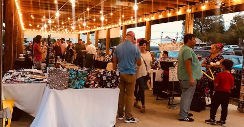 A lively market scene with people browsing stalls under string lights, showcasing various handmade items and crafts.