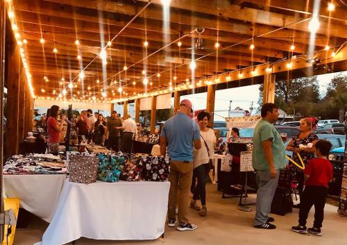 A lively outdoor market scene with people browsing tables under string lights at dusk.