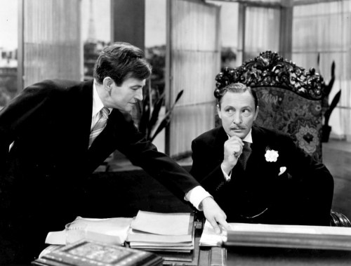 Two men in formal attire engage in a serious discussion at a desk, with papers and a large chair in the background.