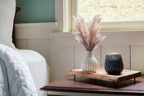 A cozy bedside table with a vase of dried flowers and a decorative candle on a wooden tray by a window.