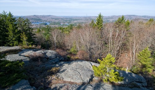 A rocky outcrop overlooks a scenic landscape of trees and distant hills under a clear blue sky.