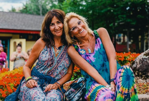 Two smiling women in colorful outfits sit together on a rock surrounded by vibrant flowers in a park setting.