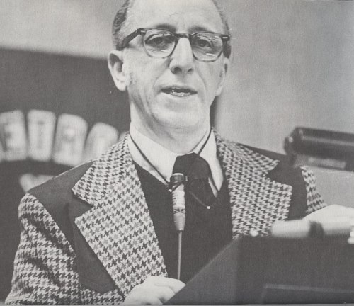 A man in a houndstooth blazer speaks at a podium, wearing glasses and a tie, with a serious expression.