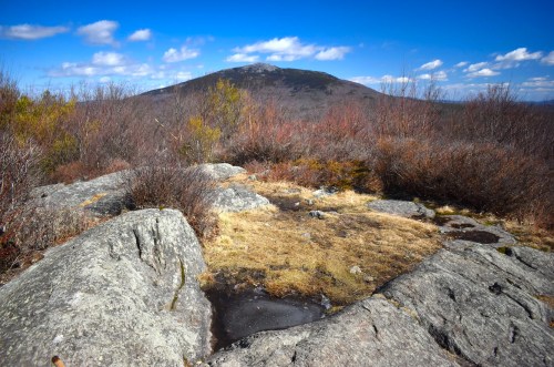 A rocky landscape with sparse vegetation, overlooking a distant mountain under a blue sky with scattered clouds.