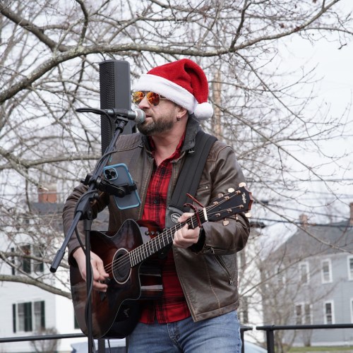 A musician in a Santa hat performs with an acoustic guitar, singing into a microphone outdoors.