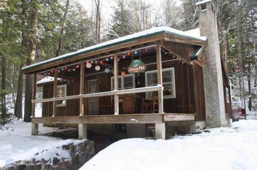 A cozy wooden cabin in a snowy landscape, adorned with colorful lights and surrounded by tall trees.