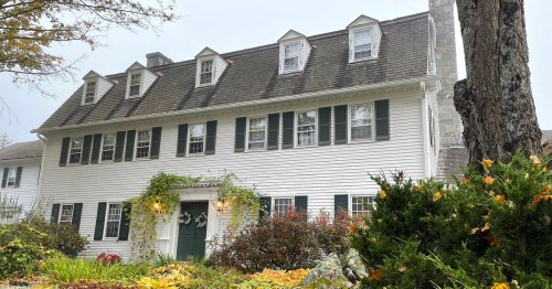 A charming two-story white house with green shutters, surrounded by colorful autumn foliage and a stone chimney.