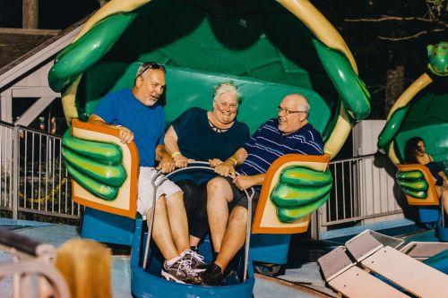 Three adults smile and sit together in a colorful amusement park ride, ready for a fun experience.