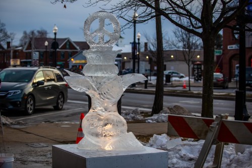 An illuminated ice sculpture featuring a peace symbol, surrounded by snow and parked cars on a city street.