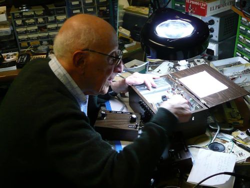An elderly man works on electronic equipment in a cluttered workshop, illuminated by a desk lamp.