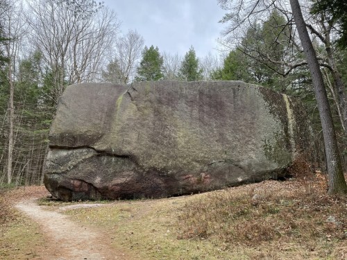 A large, flat boulder stands beside a dirt path, surrounded by trees and sparse vegetation.