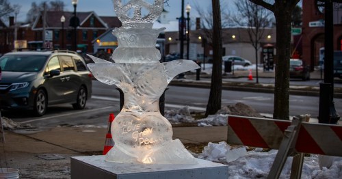 An illuminated ice sculpture shaped like a fountain, featuring a heart with the words "Love Gives Peace." Snowy street scene.