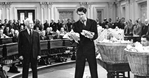 A man stands in a crowded legislative chamber, holding papers, while another man watches intently.
