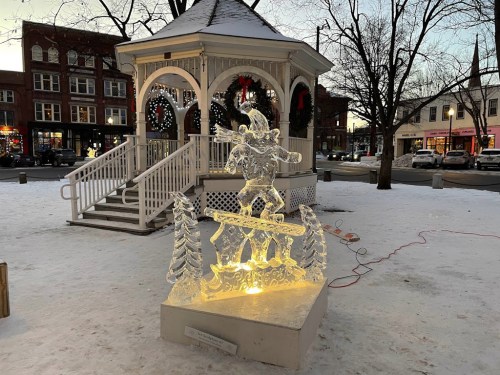 A winter scene featuring a gazebo and an illuminated ice sculpture of a figure surrounded by trees, set in a snowy park.