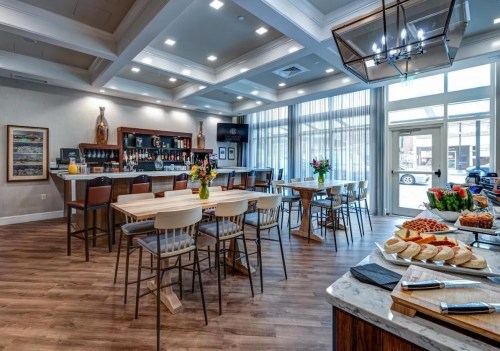 Modern dining area with wooden tables, bar, and large windows, featuring flowers and a buffet spread.