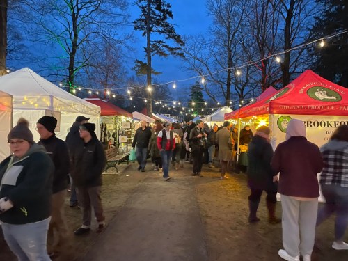 A bustling outdoor market at dusk, with colorful tents, string lights, and people browsing various stalls.