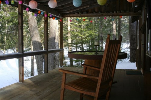 A cozy porch with a wooden rocking chair, a round table, and colorful lanterns, surrounded by snowy trees.