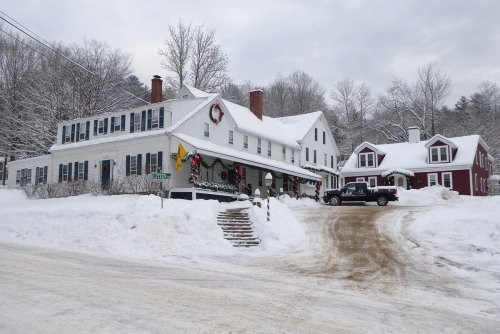 A snowy scene featuring a large white house decorated for the holidays, with a red house nearby and a truck parked on the road.