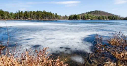A serene lake partially covered in ice, surrounded by trees and a distant hill under a clear blue sky.