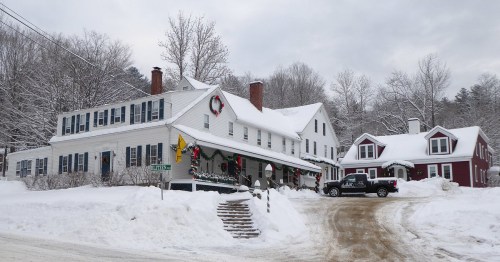 A snowy scene featuring a white house decorated for the holidays, with a red house nearby and a truck parked on a snowy road.