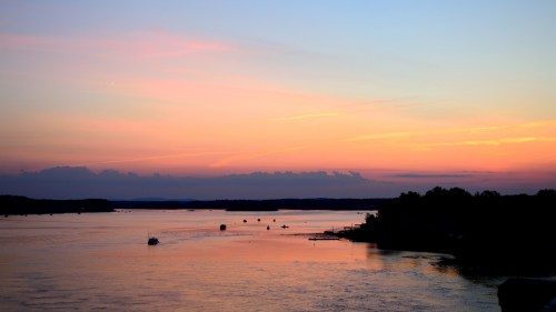 A serene sunset over a calm river, with soft pink and orange hues reflecting on the water and silhouettes of boats.
