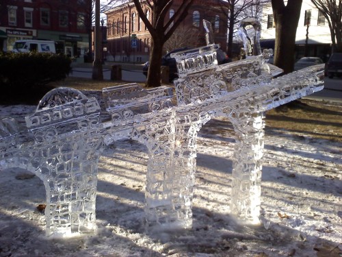 Ice sculpture of a bridge with intricate details, set in a snowy park with buildings in the background.