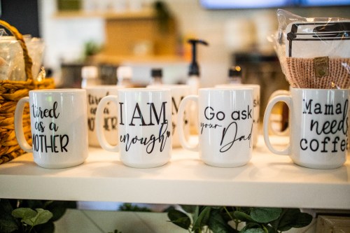 Four coffee mugs with playful phrases displayed on a shelf, surrounded by various items and greenery.