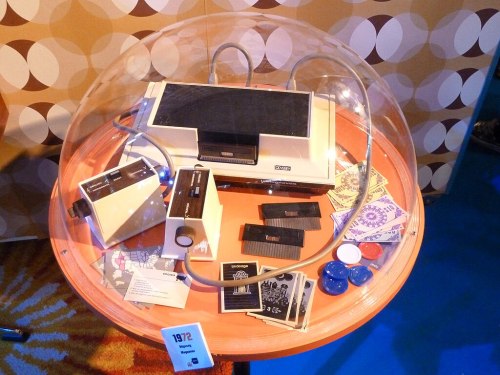 A vintage video game console displayed under a dome, surrounded by game cartridges and accessories on a circular table.